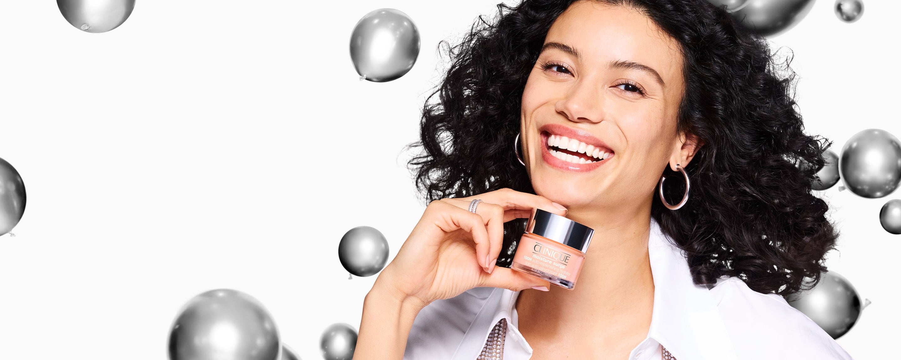 Close up of brunette model smiling, holding a Moisture Surge Moisturiser, in front of silver balloons falling in the air